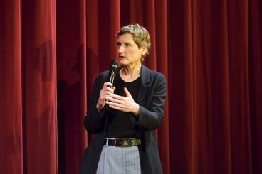 A woman with short hair is standing in front of a red curtain with a microphone in her hand, speaking. 