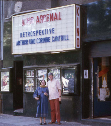 Portrait of Arthur and Corinne Cantrill in front of the program display at the old Arsenal, which reads "Retrospective Arthur and Corinne Cantrill". 