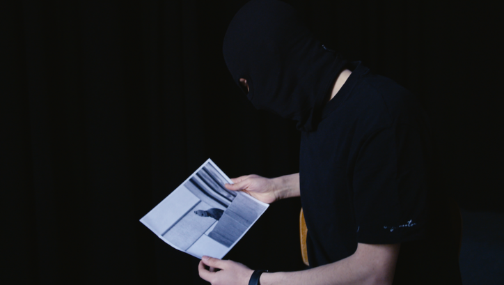 Film still from SPRICH BILD, SPRICH: A person dressed in black with a black mask over their head is holding a photo showing another person who is also wearing a mask.