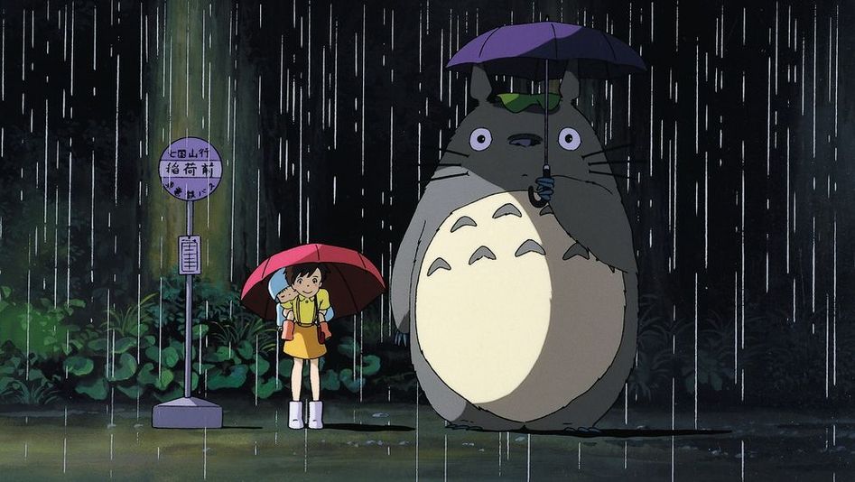Filmstill aus MEIN NACHBAR TOTORO: Zeichnung eines Mädchens, das im Regen mit einem Schirm an der Bushaltestelle steht. Neben ihr der Waldgeist Totoro mit einem großen rundlichen Körper.