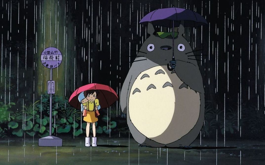 Filmstill aus MEIN NACHBAR TOTORO: Zeichnung eines Mädchens, das im Regen mit einem Schirm an der Bushaltestelle steht. Neben ihr der Waldgeist Totoro mit einem großen rundlichen Körper.