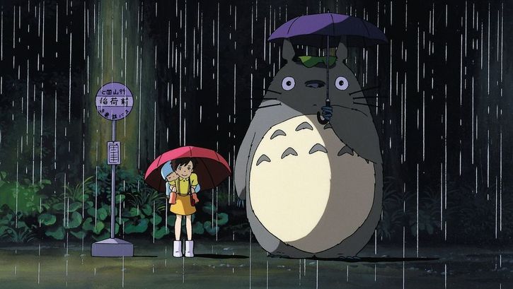 Filmstill aus MEIN NACHBAR TOTORO: Zeichnung eines Mädchens, das im Regen mit einem Schirm an der Bushaltestelle steht. Neben ihr der Waldgeist Totoro mit einem großen rundlichen Körper.