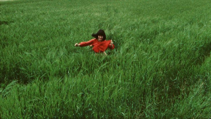 Filmstill aus GESCHICHTEN VOM KÜBELKIND: In einem Feld aus hohem, grünen Gras läuft eine Frau mit schwarzen Haaren und rotem Oberteil.