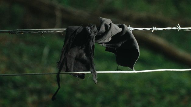 Film still from Dan Guthrie’s film "Black Strangers". A piece of cloth lodged on a horizontally spanned barbed wire.