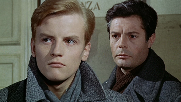 Film still from CRONACA FAMILIARE. Two men in winter coats stand one behind the other, one of them is Marcello Mastroianni.