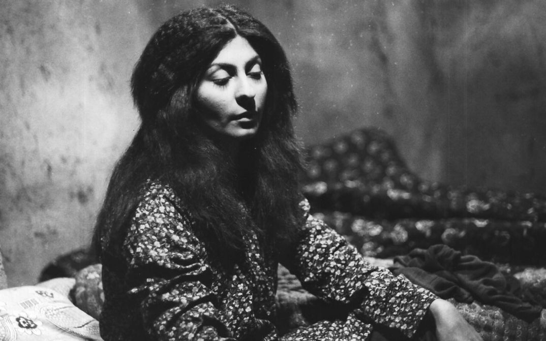 Filmstill aus THE SEALED SOIL. Eine junge Frau mit langen Haaren sitzt mit halbgeschlossenen Augen in einem Zimmer.