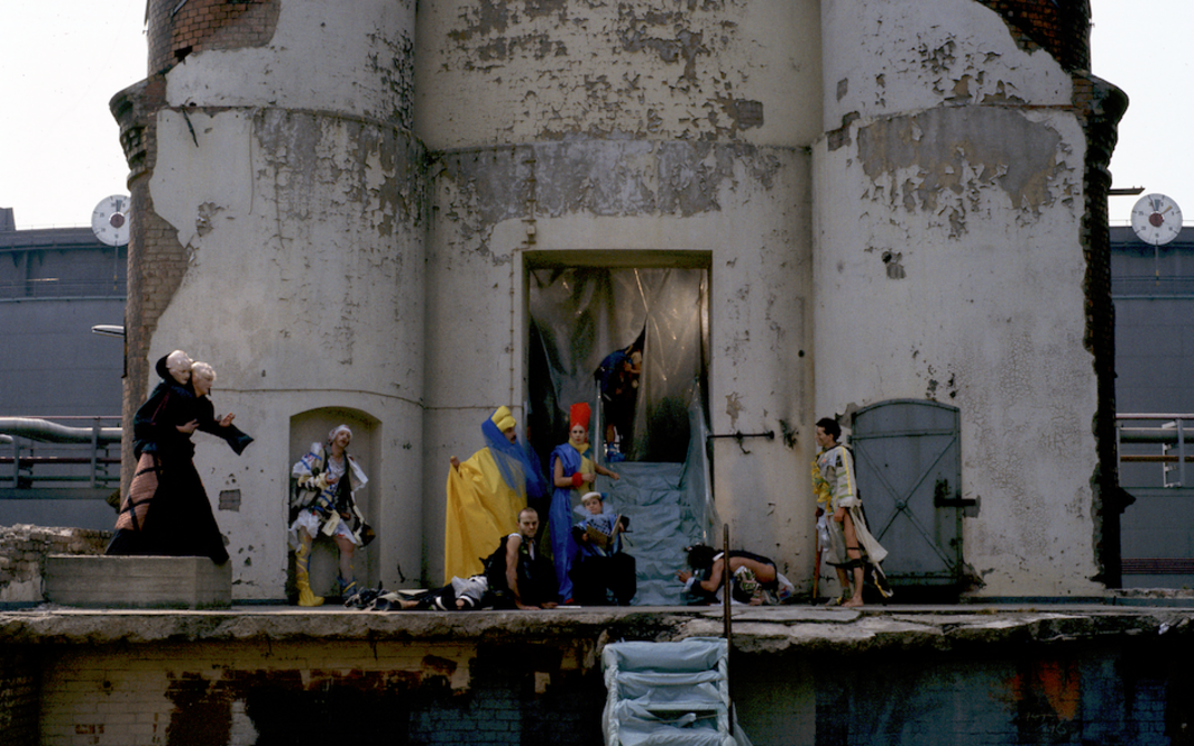 Filmstill aus USINIMAGE: Vor einem heruntergekommenen Industriegebäude sind eine Reihe von Menschen in fantasievollen Kostümen zu sehen.
