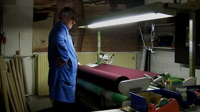 Filmstill aus „Dilim dönmüyor – Meine Zunge dreht sich nicht“ von Serpil Turhan. Ein Mann in einem blauen Arbeitskittel steht vor einer Textilmaschine. 