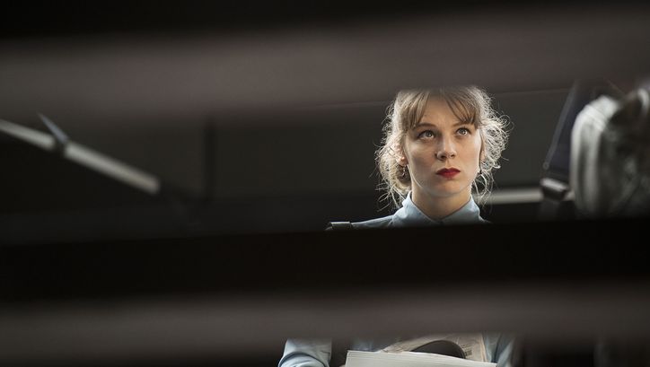 Film still from DIE LÜGEN DER SIEGER: A young woman with a serious expression is holding a piece of paper. She is visible only through a half-closed blind.