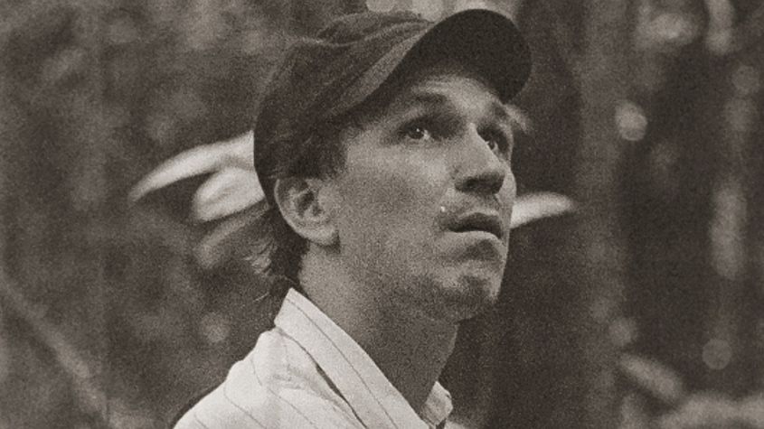 Portrait of Eduardo Williams with light shirt and cap looking up diagonally. 