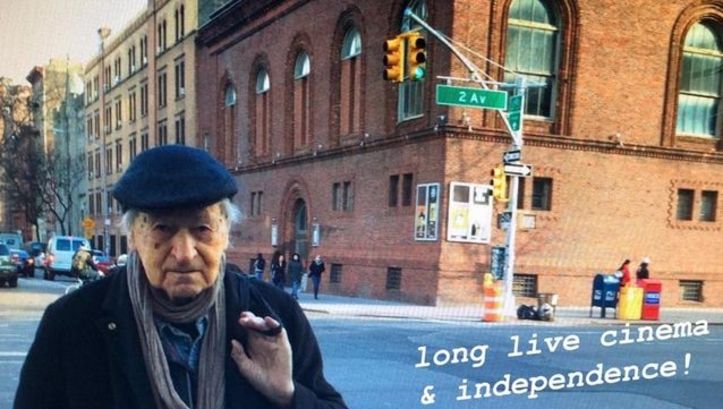 Film still from JONAS IN THE FIELDS.  Jonas Mekas is standing on a street corner in New York, with the Anthology Film Archives building in the background. On the bottom right is the writing "long live cinema & independence!".