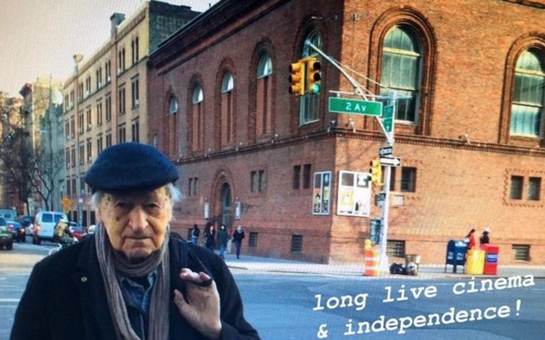 Filmstill aus JONAS IN THE FIELDS. Jonas Mekas steht in New York an einer Straßenecke, im Hintergrund das Gebäude der Anthology Film Archives. Rechts unten steht der Schriftzug "long live cinema & independence!"