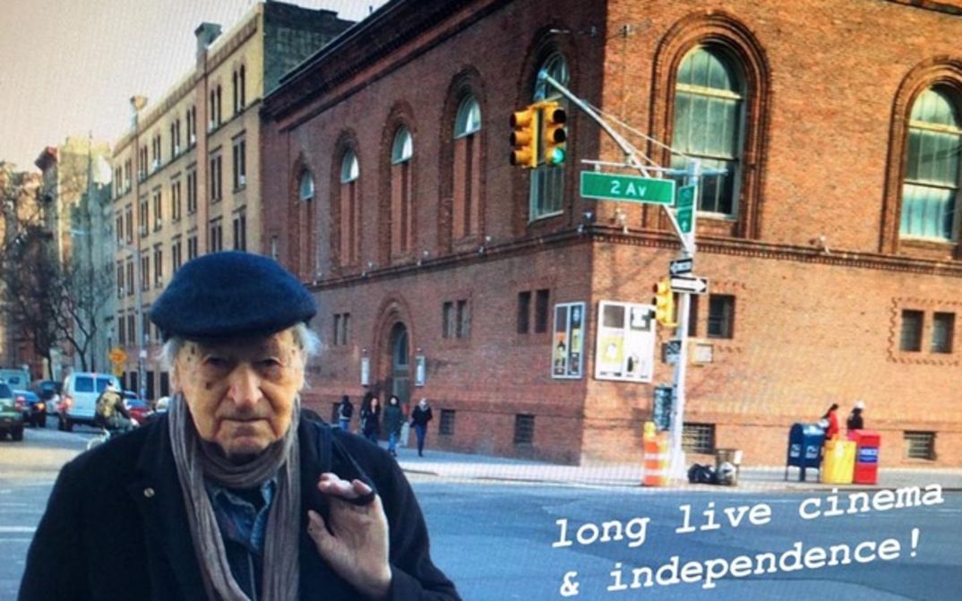 Filmstill aus JONAS IN THE FIELDS. Jonas Mekas steht in New York an einer Straßenecke, im Hintergrund das Gebäude der Anthology Film Archives. Rechts unten steht der Schriftzug "long live cinema & independence!"