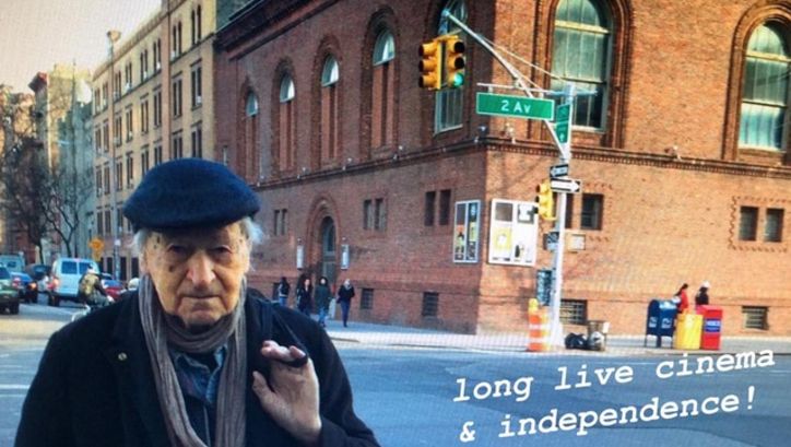 Filmstill aus JONAS IN THE FIELDS. Jonas Mekas steht in New York an einer Straßenecke, im Hintergrund das Gebäude der Anthology Film Archives. Rechts unten steht der Schriftzug "long live cinema & independence!"