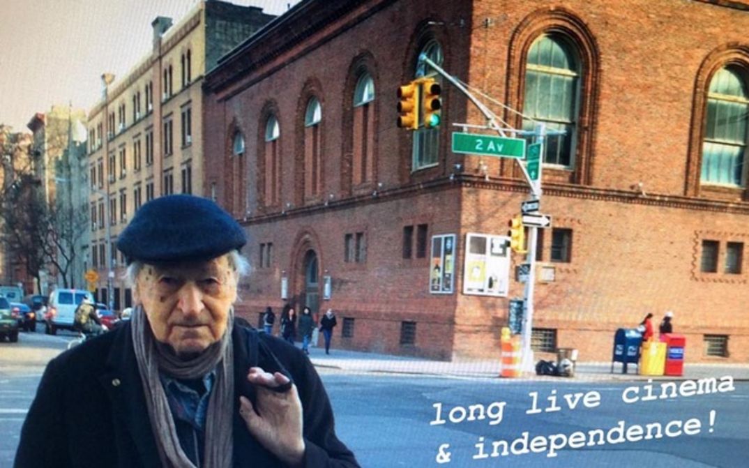 Film still from JONAS IN THE FIELDS. Jonas Mekas is standing on a street corner in New York, with the Anthology Film Archives building in the background. On the bottom right is the writing "long live cinema & independence!".