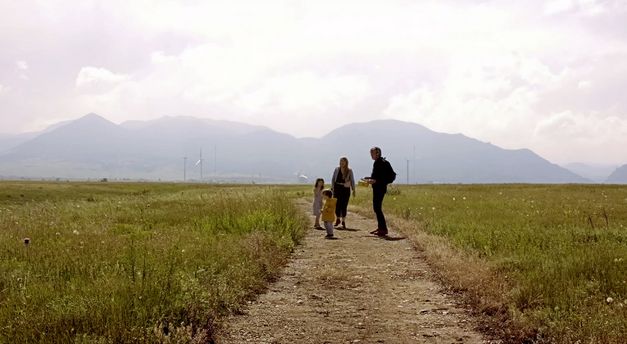 Filmstill aus „Nuclear Family“ von Erin Wilkerson und Travis Wilkerson. Zwei Erwachsene und zwei Kinder stehen auf einem Weg. Auf beiden Seiten des Weges ist Wiese. Im Hintergrund sieht man einen Berg.