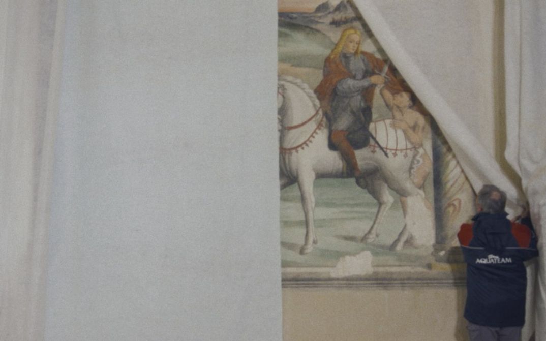 Film still from CANONE EFFIMERO by Gianluca De Serio and Massimiliano De Serio. In the picture, a man pulls back a white curtain, revealing a painting behind it.