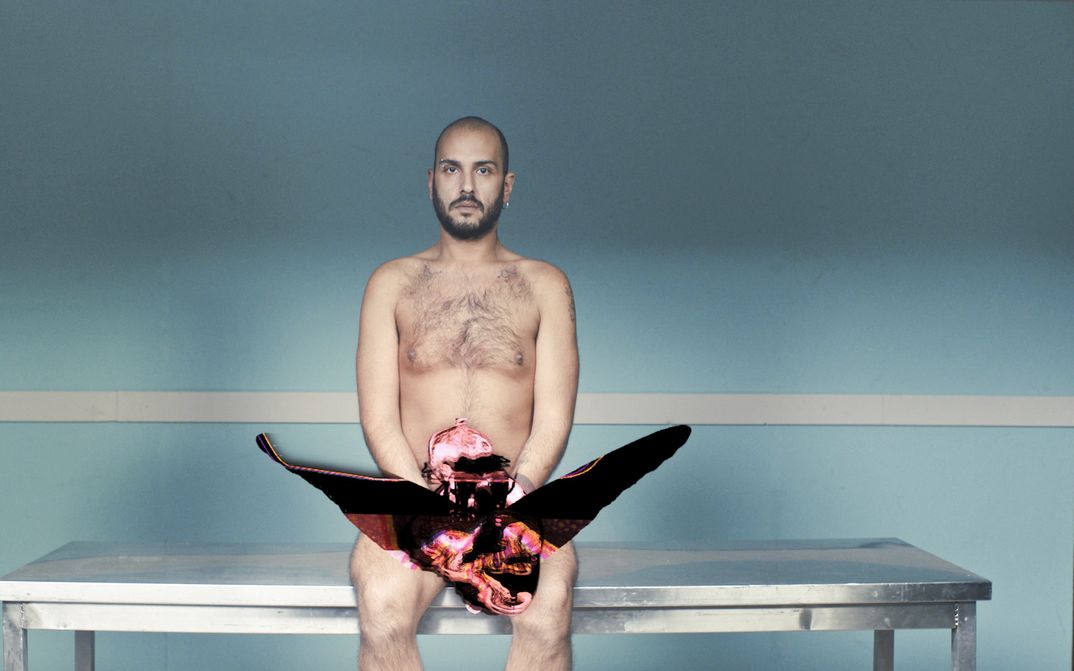 Film still from EMBODIED CHORUS: A young man sits naked on a bench in front of a turquoise-painted wall.