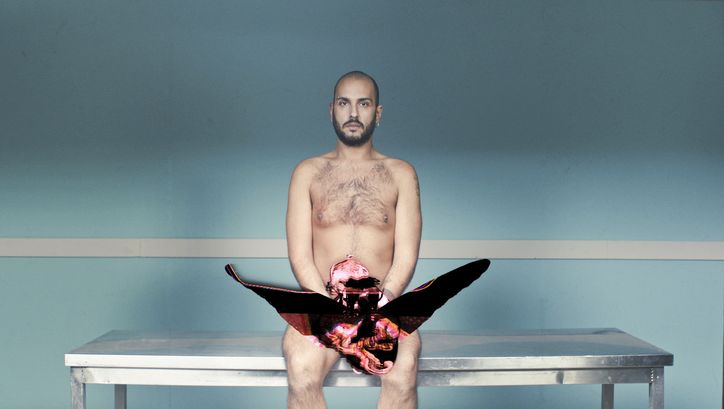Film still from EMBODIED CHORUS: A young man sits naked on a bench in front of a turquoise-painted wall. 