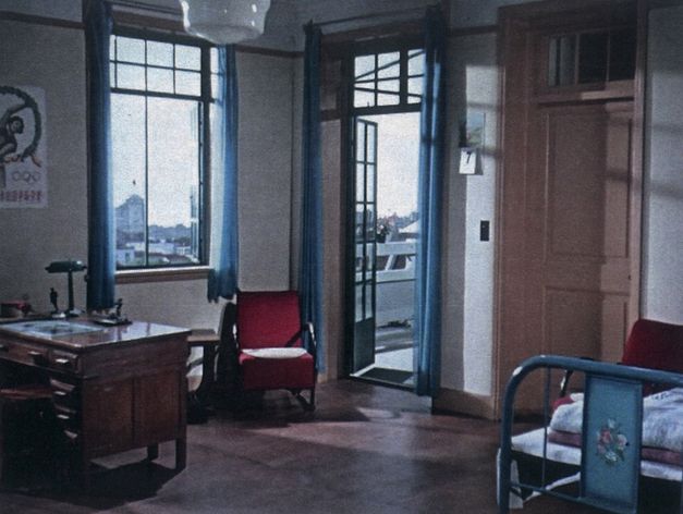 Film still from Lei Lei’s „That Day, on the River“. The interior view of an apartment with a bed, desk, chair and curtains. Through the windows and the open balcony door we see a city.