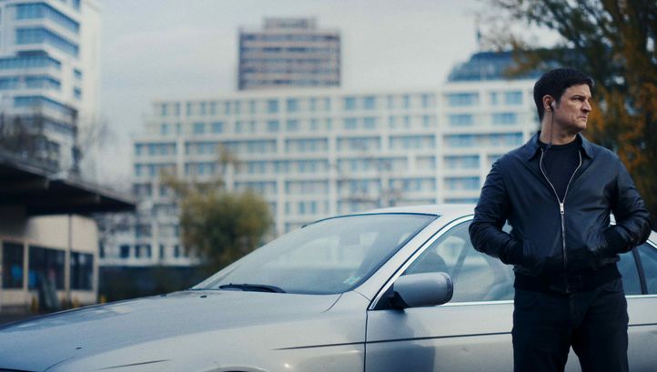 Filmstill aus VERBRANNTE ERDE: Ein Mann steht mit den Händen in den Taschen vor einem silbernen Auto. Er hat Kopfhörer im Ohr. Im Hintergrund sind Hochhäuser.