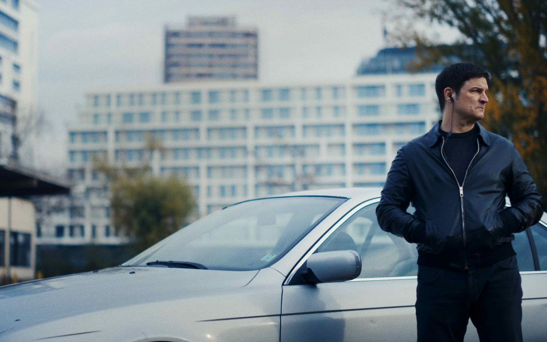 Film still from VERBRANNTE ERDE: A man stands in front of a silver car with his hands in his pockets. He has headphones in his ears. There are tower blocks in the background.