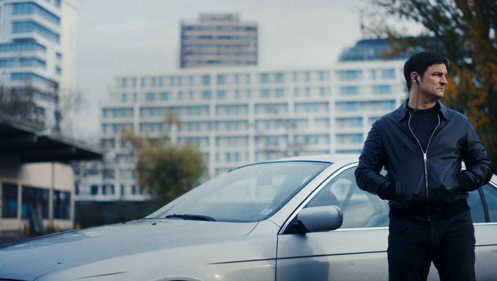 Filmstill aus VERBRANNTE ERDE: Ein Mann steht mit den Händen in den Taschen vor einem silbernen Auto. Er hat Kopfhörer im Ohr. Im Hintergrund sind Hochhäuser.