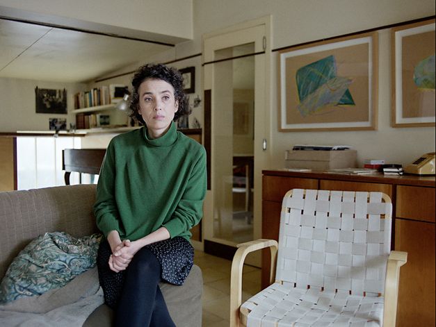 Still from the film "O trio em mi bemol (The Kegelstatt Trio)" by Rita Azevedo Gomes. A woman in a green sweater sits looking pensively with her legs crossed on the end of a sofa.  