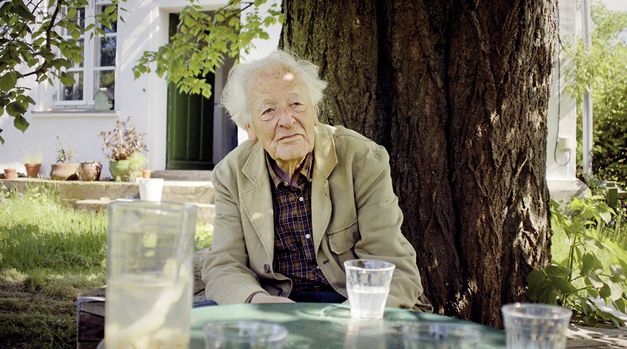 Still from the film "Gehen und Bleiben" by Volker Koepp. An elderly man with a checkered shirt and a beige jacket is sitting at a table in front of a tree in a garden. Several glasses standing on the table are blurred out in the foreground. In the background, a dark green door leading to a house is partially open. 