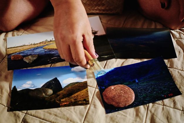 Still from the film "O Estranho" by Flora Dias and Juruna Mallon. Five coloured photographs are placed on top of a blanket, there are grey and red stones on top of them. There is a hand with dark varnish on the fingernails hovering above them, holding a lighter.