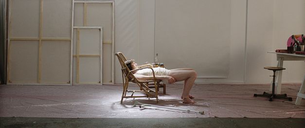 Filmstill aus "Mit einem Tiger schlafen" von Anja Salomonowitz. Zu sehen ist eine Person in Unterwäsche, die halb auf einem Stuhl in der Mitte eines Raums im Scheinwerferlicht liegt. Im Hintergrund befinden sich Leinwände an der Wand. Rechts im Bild steht ein Schreibtisch mit Malutensilien 