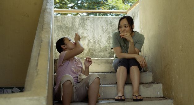 Filmstill aus "Oasis of Now" von Chee Sum Chia. Zu sehen sind eine Frau und ein Kind auf einer Betontreppe. Im Hintergrund scheint draußen die Sonne. 