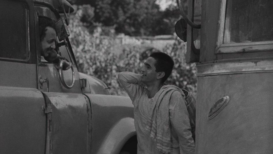 Film still from BADNAM BASTI: A young man is standing on the street between cars, talking to a man who is looking out the window of a truck.