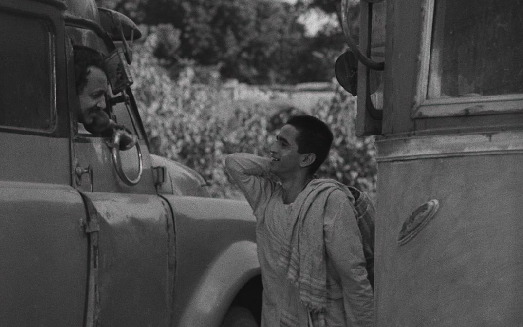 Filmstill aus BADNAM BASTI: Ein junger Mann steht auf der Straße zwischen Autos und unterhält sich mit einem Mann, der aus dem Fenster eines LKWs blickt.