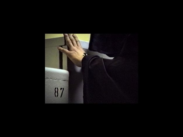 Film still from Anna Zett’s film “Es gibt keine Angst (Afraid Doesn’t Exist)”. On the right a person in a black robe putting their left hand on a shelf on the left. In the bottom left corner another shelf with the number 87 on its door.