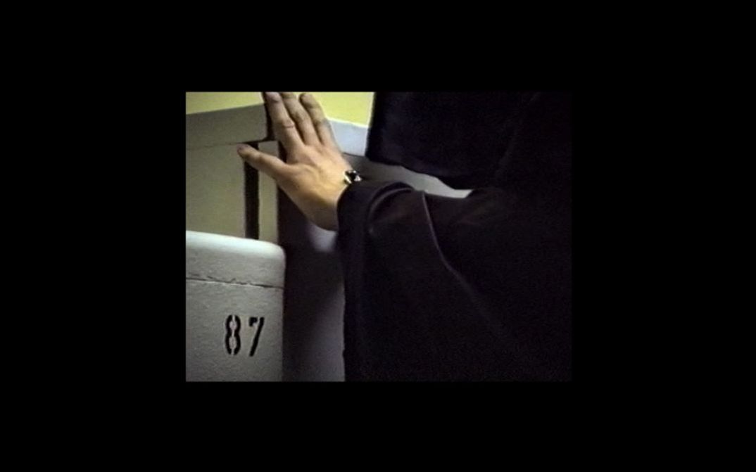 Filmstill aus dem Film „Es gibt keine Angst“ von Anna Zett. Eine Person in schwarzer Kutte auf der rechten Seite, die eine Hand auf einen Schrank auf der linken Seite legt. Auf einer weiteren Schranktür in der linken unteren Ecke die Nummer 87.