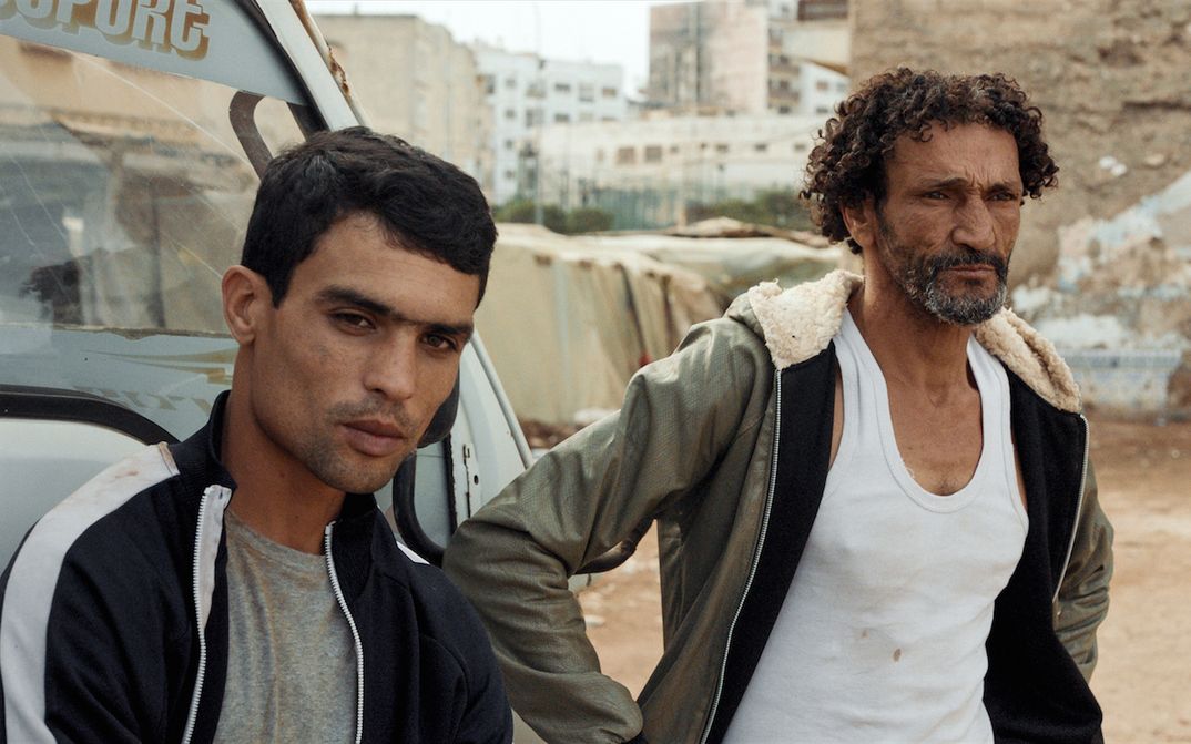 Film still from HOUNDS: Two men in simple clothes are standing next to a car in a city.