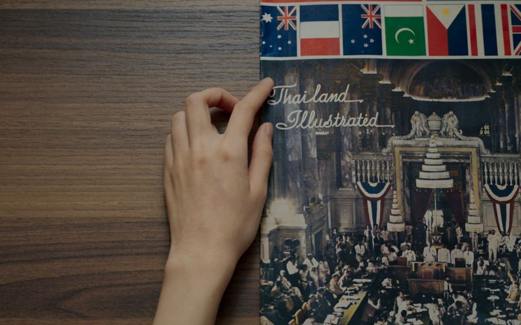 Film still from Chanasorn Chaikitiporn’s film “Here We Are”. A hand traces a book on a wooden table titled “Thailand Illustrated”.