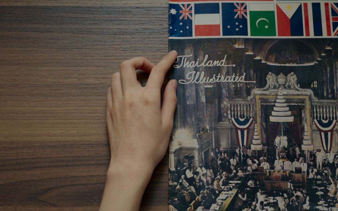 Film still from Chanasorn Chaikitiporn’s film “Here We Are”. A hand traces a book on a wooden table titled “Thailand Illustrated”.