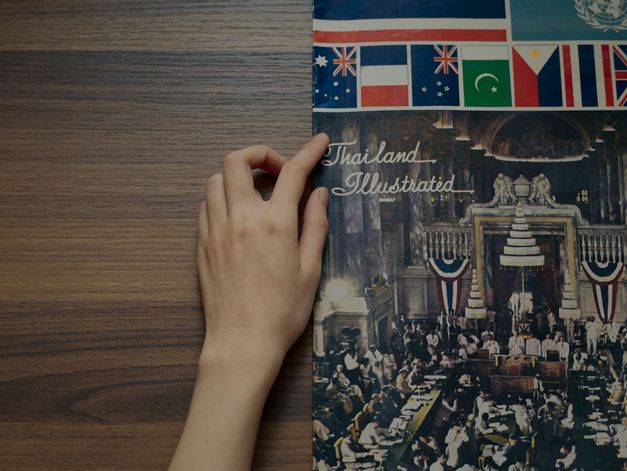 Filmstill aus dem Film „Here We Are“ von Chanasorn Chaikitiporn. ”. Eine Hand zeichnet ein Buch auf einem Holztisch mit dem Titel "Thailand Illustrated".	