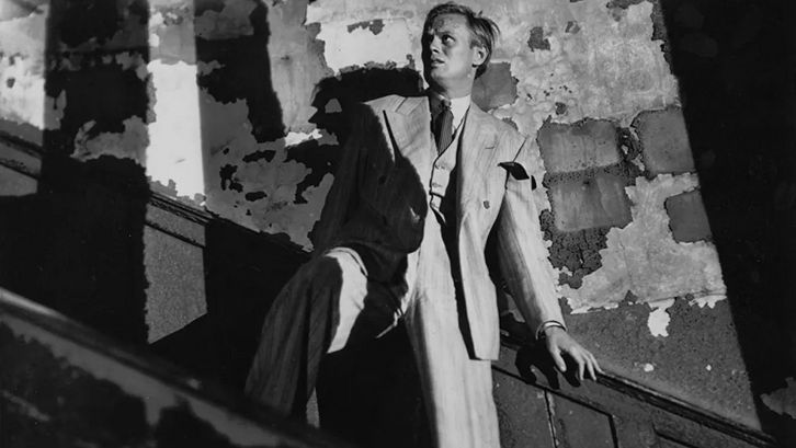 Filmstill aus NIGHT AND THE CITY: In einem heruntergekommenen Treppenhaus hält sich ein Mann am Geländer fest und blickt mit verängstigtem Gesichtsausdruck nach oben.