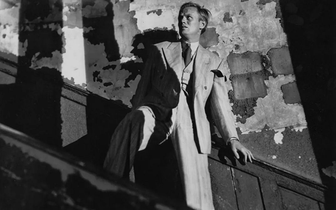 Filmstill aus NIGHT AND THE CITY: In einem heruntergekommenen Treppenhaus hält sich ein Mann am Geländer fest und blickt mit verängstigtem Gesichtsausdruck nach oben.