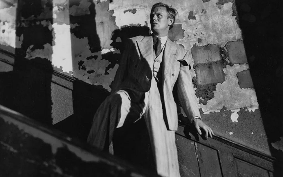 Film still from NIGHT AND THE CITY: In a dilapidated stairwell, a man is holding onto the railing and looking up with a frightened expression on his face.