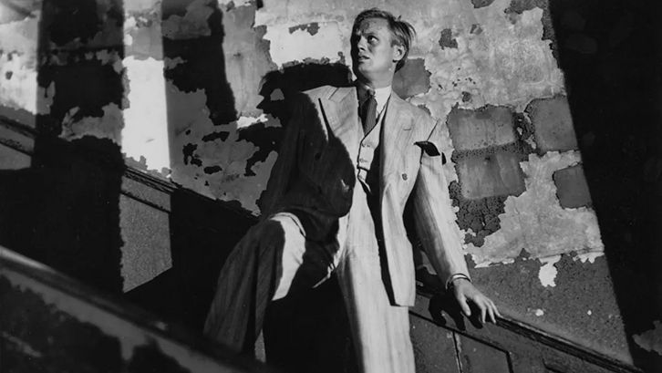 Film still from NIGHT AND THE CITY: In a dilapidated stairwell, a man is holding onto the railing and looking up with a frightened expression on his face.