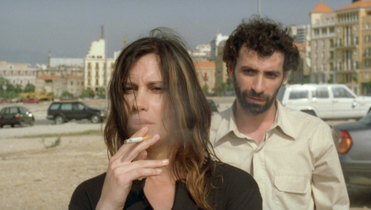 Filmstill aus TERRA INCOGNITA. Auf einer Straße steht rauchend eine Frau, hinter ihr steht ein Mann.