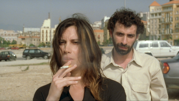 Filmstill aus TERRA INCOGNITA. Auf einer Straße steht rauchend eine Frau, hinter ihr steht ein Mann.