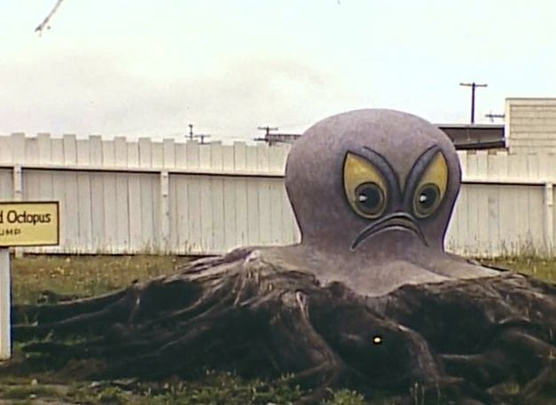 Film still from TERRA FEMME. You can see a huge octopus figure with scowling face on a meadow.