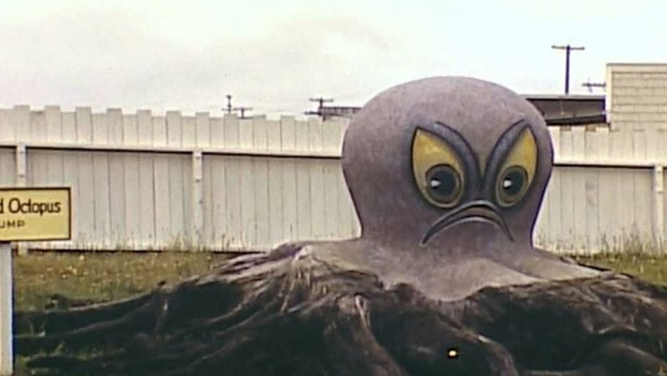 Film still from TERRA FEMME. You can see a huge octopus figure with scowling face on a meadow.