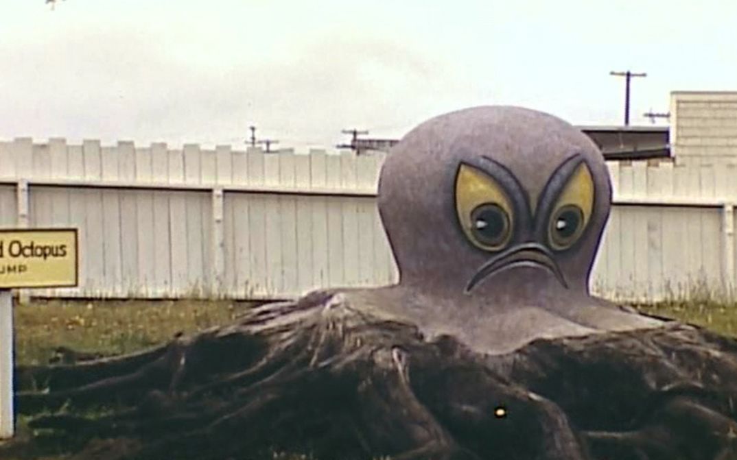 Filmstill aus TERRA FEMME. Man sieht eine riesige Oktopus-Figur mit missmutigem Gesicht auf einer Wiese.