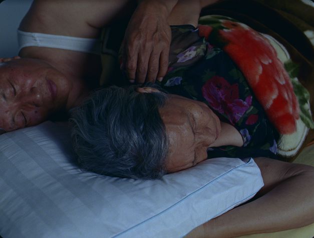 Film still from Tenzin Phuntsog’s „Dreams“. Two elderly people lying on a matress, the hand of the one’s in the back on the shoulder of the one’s in front.